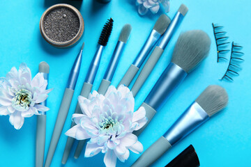 Makeup brushes with false eyelashes and flowers on blue background, closeup