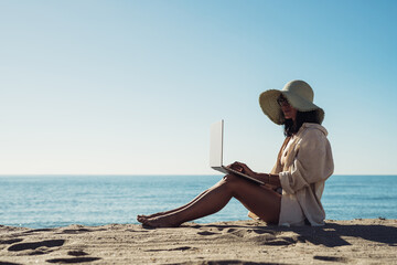 young beautiful girl freelancer with a laptop works on the seashore sitting on the sand