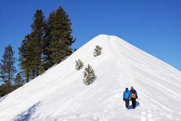 Two people exploring the Northwest Wilderness walking up a steep snow covered hill.
