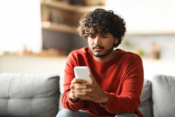 Pensive Young Indian Man Looking At Smartphone Screen While Sitting At Home