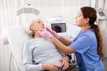 Mature woman lying on couch during consultation with female beautician, checking her skin before...