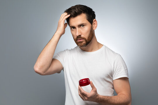 Hair care concept. Confident bearded man holding hair gel and touching his ideal hair, grey studio background