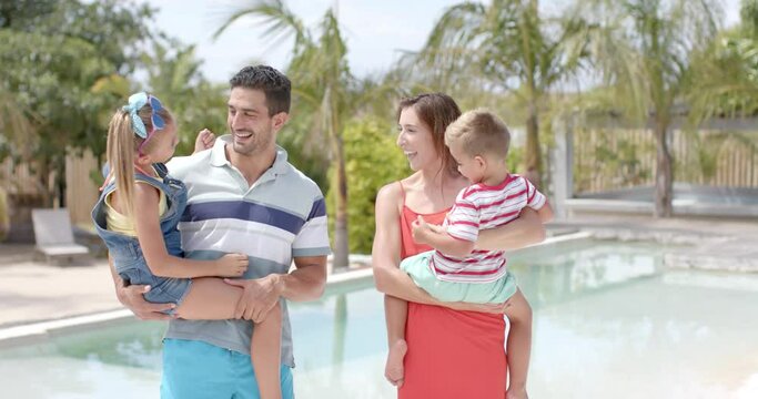 Portrait of happy caucasian parents carrying children at swimming pool at beach house