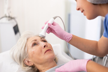 Delighted old woman lying on clinical chair in aesthetic cabinet during face laser therapy procedure