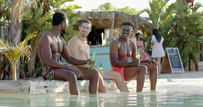 Happy diverse male friends drinking beer and talking at beach bar and pool