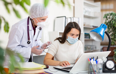 Focused business woman in protective mask sitting at workplace in office, getting vaccinated by elderly female doctor. Immunization and disease prevention concept