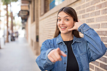 young pretty woman smiling cheerfully and pointing to camera while making a call you later gesture, talking on phone