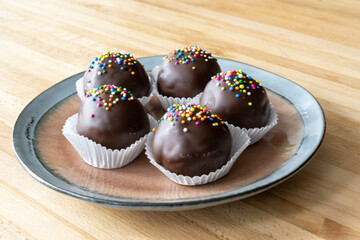 A small dinner plate with multiple chocolate covered cakes pops on a wooden table. The dark chocolate balls have multi colored sprinkles on top and are in round white parchment paper cup holders.
