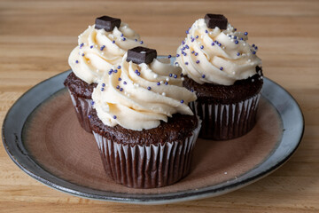 Three chocolate cupcakes with vanilla icing, sprinkles, and chocolate square on a small plate and wooden table. The homemade baked and frosted brownie style cakes swirl in buttercream icing. 