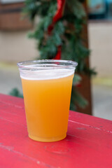 A tall clear plastic glass of sour craft beer in a microbrewery. The cold refreshment has frost on the glass. The beer is on a wooden patio table. The top of the glass has white froth around the rim. 