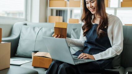Using Laptop computer, Young pretty Asian female influencer small businesses SME owners female entrepreneurs working on receipt box and check online orders, sitting on sofa of home online business.