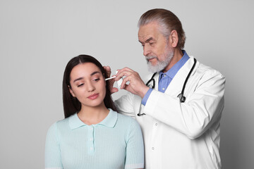 Doctor spraying medication into woman's ear on light grey background
