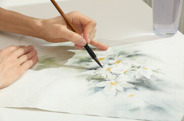 Woman painting flowers with watercolor at white table, closeup. Creative artwork