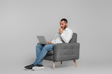 Thoughtful young man with laptop sitting in armchair on grey background