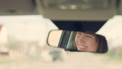 Beautiful young woman traveling by car, smiling, rejoicing, good mood. Young female driver in reflection of rearview mirror. Girl driver drives her car, happy emotions. Weekend, car, road, sports