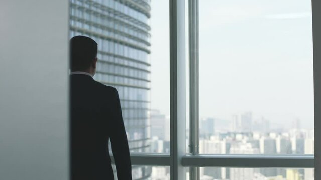 rear view of an asian business man walking up to the window looking at city through glass