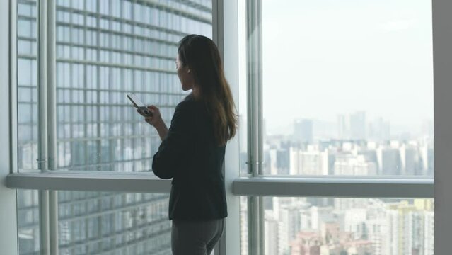 young asian businesswoman looking at cellphone while pacing by the window in silhouette
