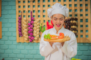 Beautiful asian female professional chef holding and looking to a wooden tray with tomatoes fresh green vegetables and bread in the kitchen full of cooking ingredients.