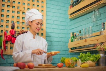 Beautiful asian female professional chef spread the sauce on the bread with joyful in the kitchen full of cooking ingredients.