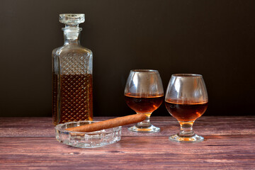 A Cuban cigar in a glass ashtray, a crystal decanter and two glasses of whiskey on a dark wooden table.