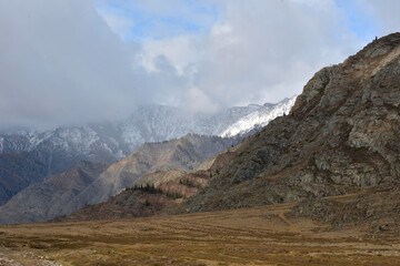 Steppe at the foot of a high mountain range with snow-capped peaks in the shadow of a thunderstorm front.