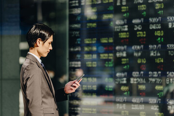 株価ボードの映る電光掲示板を前にスマートフォンで株を売買する30代のビジネスマン投資家