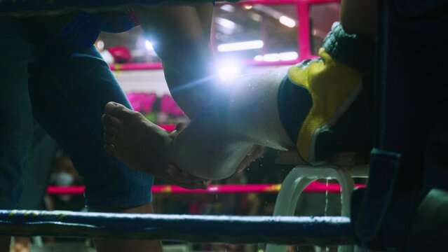 4K Cinematic slow motion footage of a Muay Thai coach pouring water on his Thai fighter and massaging his leg on a boxing ring during a Muay Thai boxing fight in a stadium