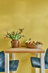 two chairs and a table with flowers in the middle, on a yellow wall behind it is a basket of fruit