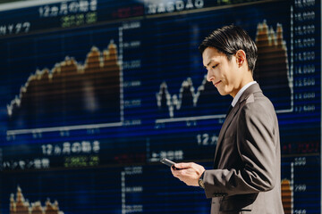 株価ボードと株価チャートのうつる電光掲示板の前でスマートフォンを見る30代の日本人の男性