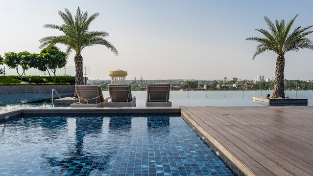 Infinity pool on the roof of the building. The blue tiled bottom is visible through the clear water. Boardwalk. The deck chairs are in a row. Ahead, behind a transparent fence, the city is visible. 