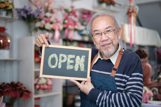 One senior male florist owner in apron shows open sign board in bright flower shop store with smile and looks at camera, small business opening, occupation retirement, happy elderly SME entrepreneur.