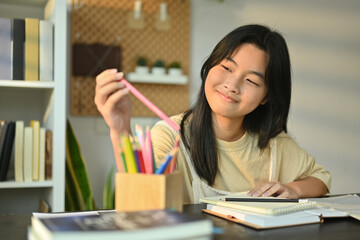 Cheerful teenaged girl doing homework, writing in notebook while sitting at table in living room