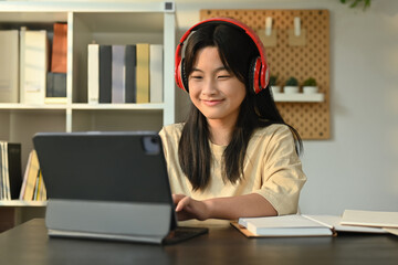 Happy asian girl wearing headphones sitting at table learning online at virtual class on laptop....