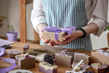 Woman in an apron holds a craft box with a ribbon in her hands. Lavender flowers and natural soap...