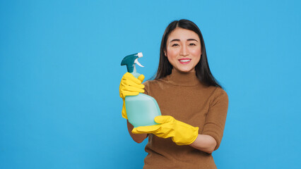 Smiling asian maid wearing protective gloves while showing chemical detergent spray in front of camera, promoting cleaning product. Professional housekepper worked hard to maintain a clean home