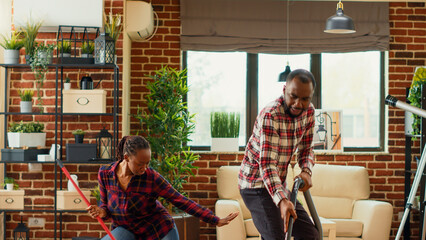 Happy husband and wife enjoying cleaning with mop and vacuum cleaner, singing and listening to music at home. Young partners mopping and vacuuming tiles in apartment. Tripod shot.