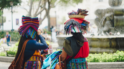 Two women street vendors talking, carrying things on their heads and selling traditional fabrics, toys and typical Guatemalan handicrafts in the Central Park of Antigua, in front of the fountain
