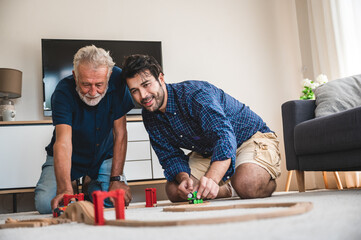 Closeup of a young man and elderly father playing with toy train set at home by sitting on floor in...