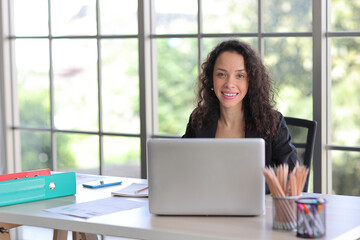 Attractive professional latin female employee worker sitting, using laptop computer with smart mobile phone at home workplace. Businesswoman working on paperwork while looking at camera and smile