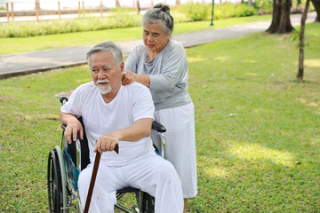 Fototapeta premium Asian senior woman caregiver helping and consoling senior man walk with wheelchair and wooden stick at park outdoor. Elderly wife taking good help care and support of elder husband patient outdoor