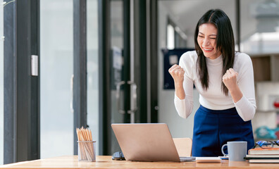 Excited asian businesswoman raising hands to congratulate while working on laptop in office.