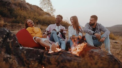 Young group of people is sitting in the dusk by the campfire, drinking beer and clinking bottles. Multiracial people have fun and sing along with the guitar outdoors on a glamping