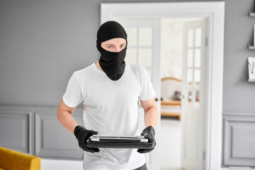 Thief with black balaclava stealing modern expensive laptop from apartment. Masked face. Man...
