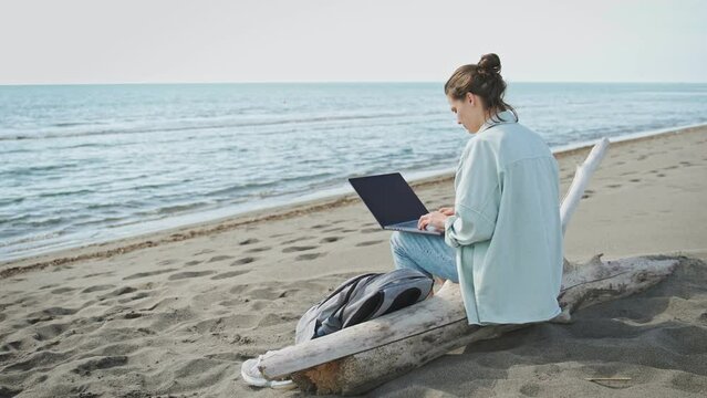 A young Caucasian woman freelancer working on a laptop while sitting on a sandy beach.