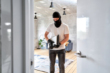 Masked face. Man burglar stealing tv set from house. Thief with black balaclava stealing modern expensive laptop from apartment.  - obrazy, fototapety, plakaty