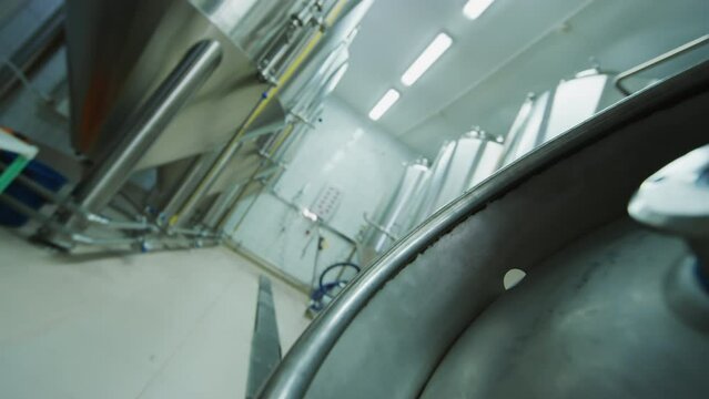 Tilt shot of beer keg on floor in brewery, legs of anonymous male technician in white coat walking up, lifting and carrying it along rows of steel fermentation tanks, then setting it down on floor