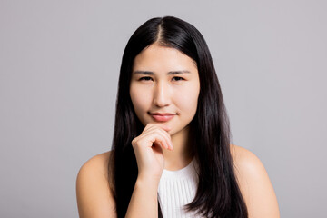 Fototapeta premium Curious thoughtful young happy asian woman with black dark long hair holding hand near face natural make up skincare welness on greu background isolated.