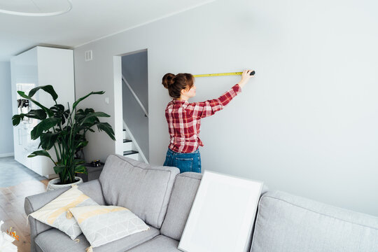 Young woman in plaid shirt doing measuring with a measure tape on the wall. Girl wants to put a picture on the wall at home. Housekeeping work. Doing repair herself. DIY, equality in work concept.