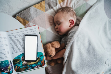 Little boy child sleeping on bed bedside table with open fairytale book and smartphone on it, kid...