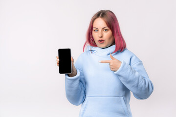 Surprised lovely woman with pink hair in a blue hoodie shows a mobile phone with a blank screen. Young female looking amazed by promotion offer, stands over white background